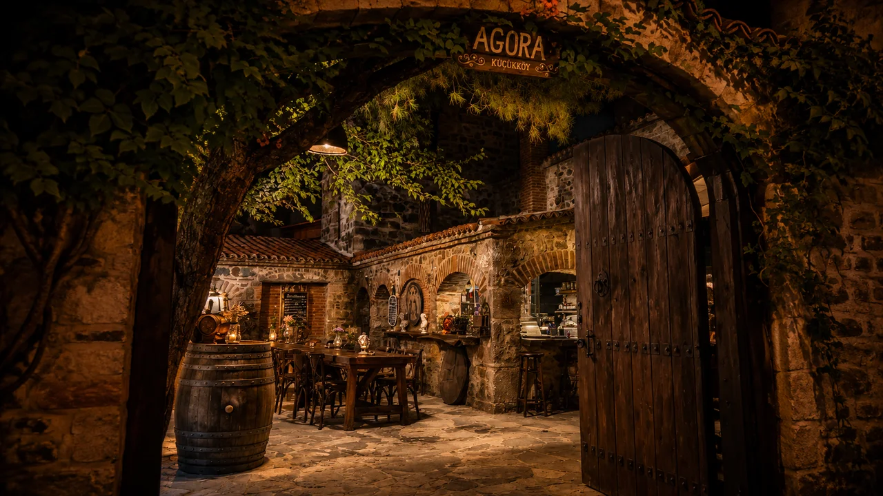 Ayvalık Küçükköy'deki Agora'nın taş kemerli girişinden görülen tarihi taş avlu, ahşap kapı, sıcak gece ışıkları ve otantik Ege atmosferi.